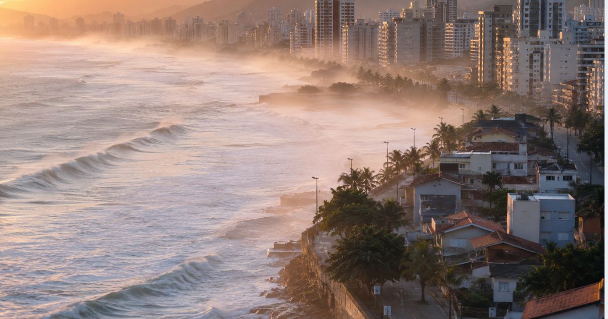 Paisagem litorânea ao entardecer com ondas quebrando e névoa salina sobre as casas, simbolizando a maresia que atinge as estruturas de esgoto.