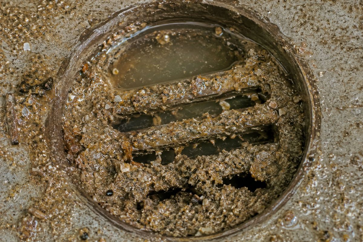 Close-up de um ralo entupido por areia e detritos, com a água acumulada.